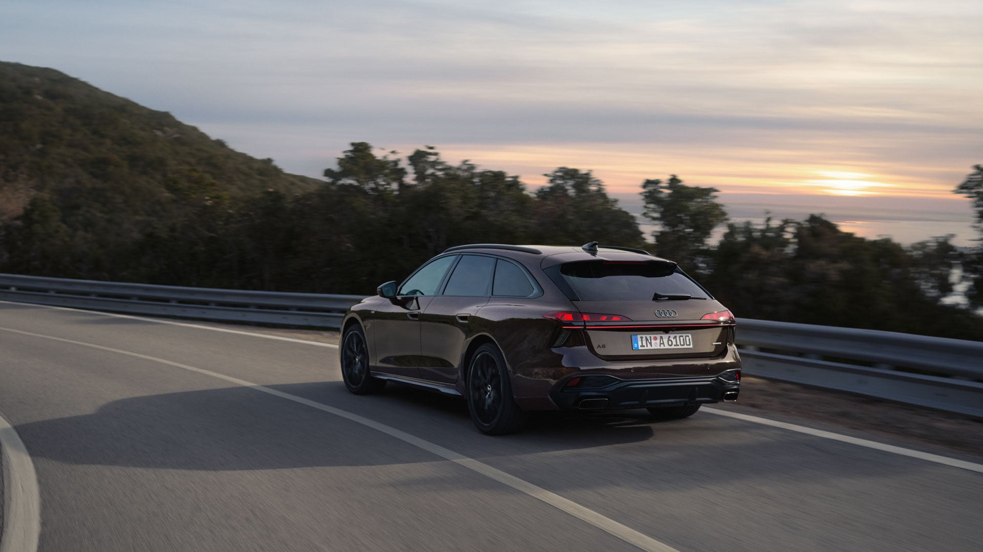 Dunkler Audi-Kombi fährt bei Sonnenuntergang auf einer kurvigen Straße entlang.