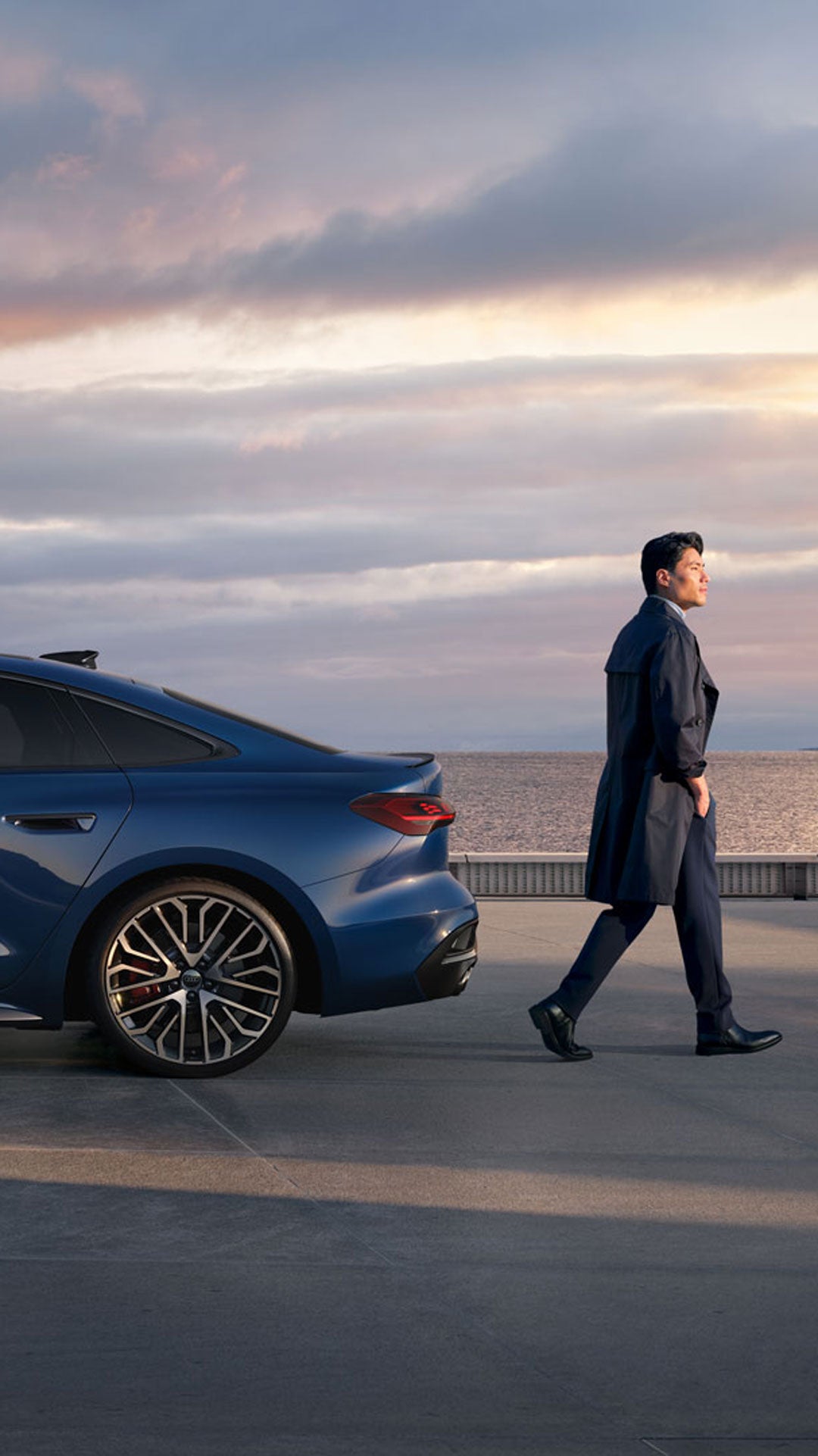 Ein Mann in dunkler Kleidung geht auf einer Promenade neben einem blauen Audi S5 vor Meer und Sonnenuntergang entlang.