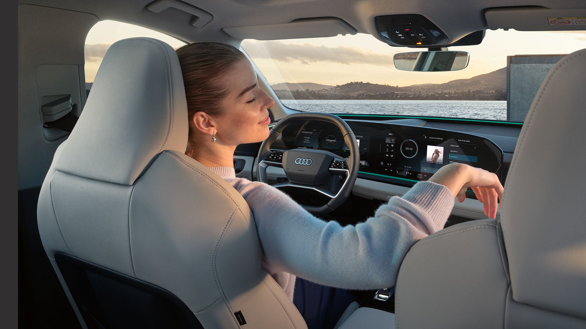A woman sat in the drivers seat in an Audi vehicle which also shows a stylish dashboard, large display screens, and scenic views through the windshield.