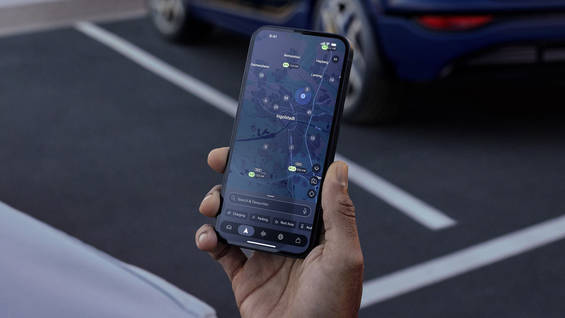 Hand holding a smartphone with a charging point search app, with the rear of an Audi vehicle in the background.
