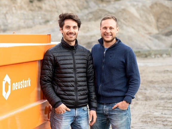 Two men stand next to a container with the inscription neustark.