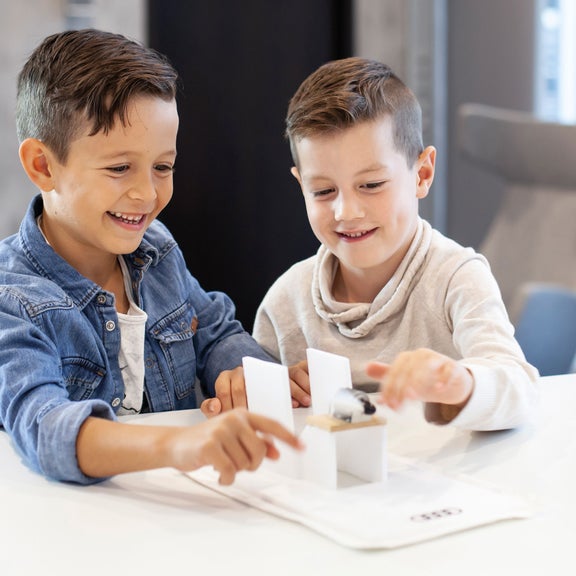 Two smiling boys playing with a small model car on a white table in a bright room.