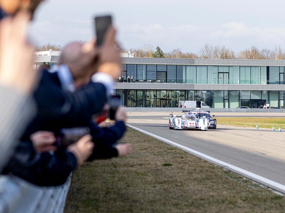 A picture showing two race cars from Audi Sport racing legends being demonstrated on the track in Neuburg, together with potential customers watching the cars.