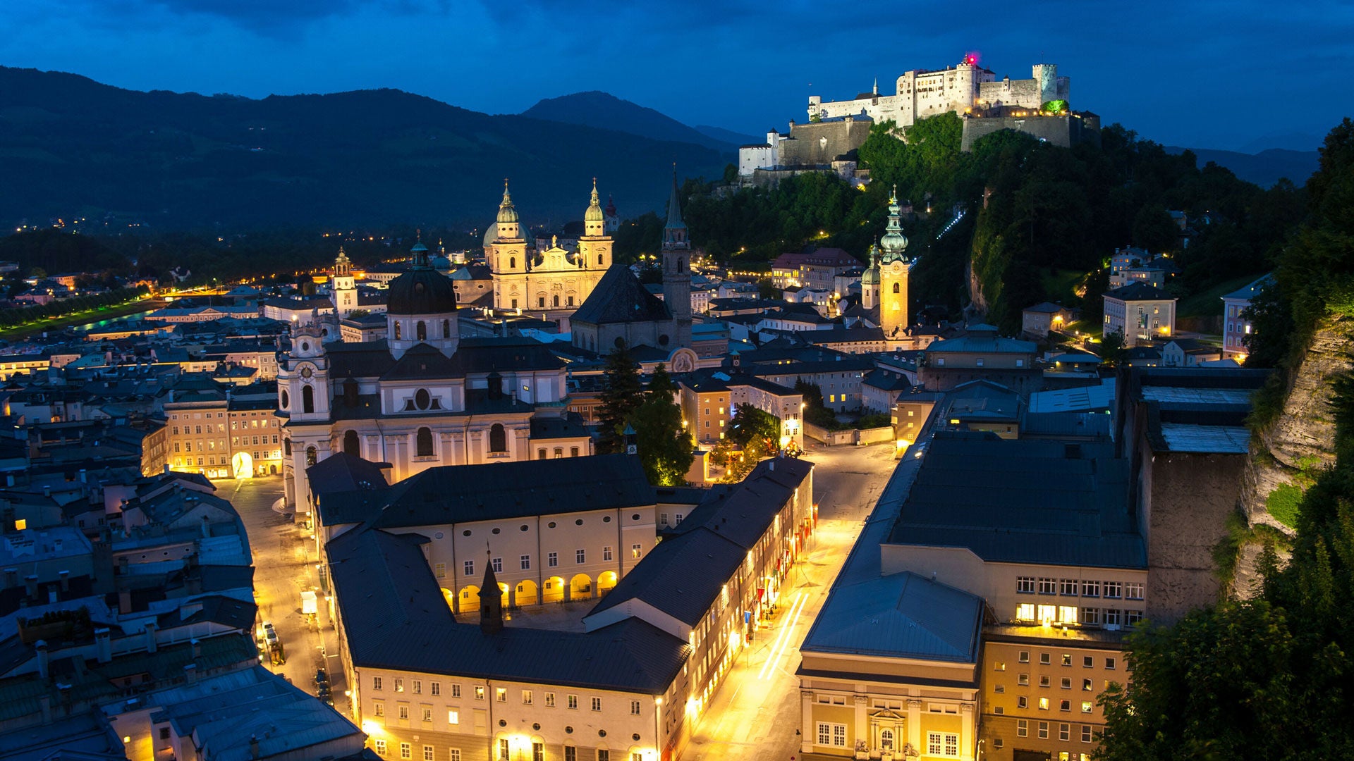 Evening city view of Salzburg with illuminated old town and Hohensalzburg Fortress in the background.