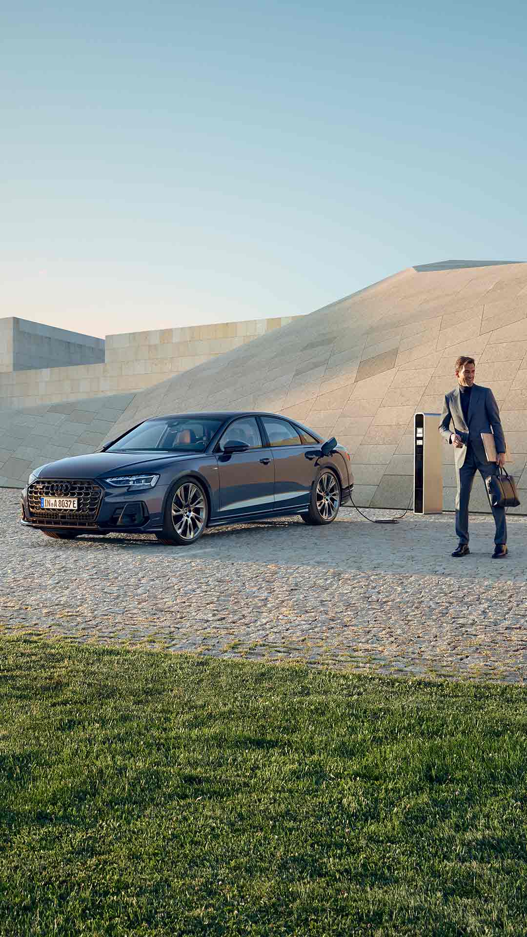 Ein dunkelblauer Audi A8 60 TFSI e lädt an einer Ladesäule, ein Mann in grauem Anzug steht mit Aktentasche und Laptop in der Hand daneben.