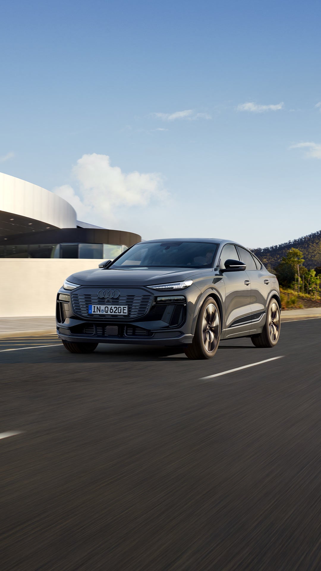 Dunkelgrauer Audi Q6 Sportback e-tron fährt auf leerer Straße vor moderner Architektur und bewaldeter Hügellandschaft.