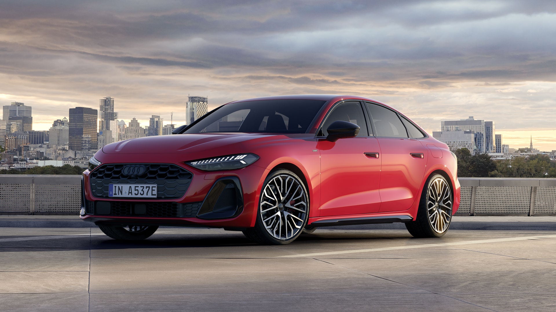 Die A5 Limousine e-hybrid im schrägen Frontprofil. Sie ist in misanorot lackiert. Im Hintergrund ist die Skyline einer Stadt bei Dämmerung zu sehen.