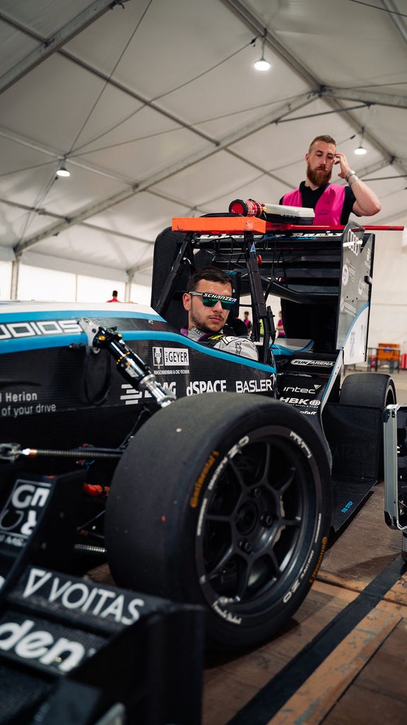 Eine Person sitzt mit Sonnenbrille auf in einem Rennauto und eine andere Person steht hinter dem Auto