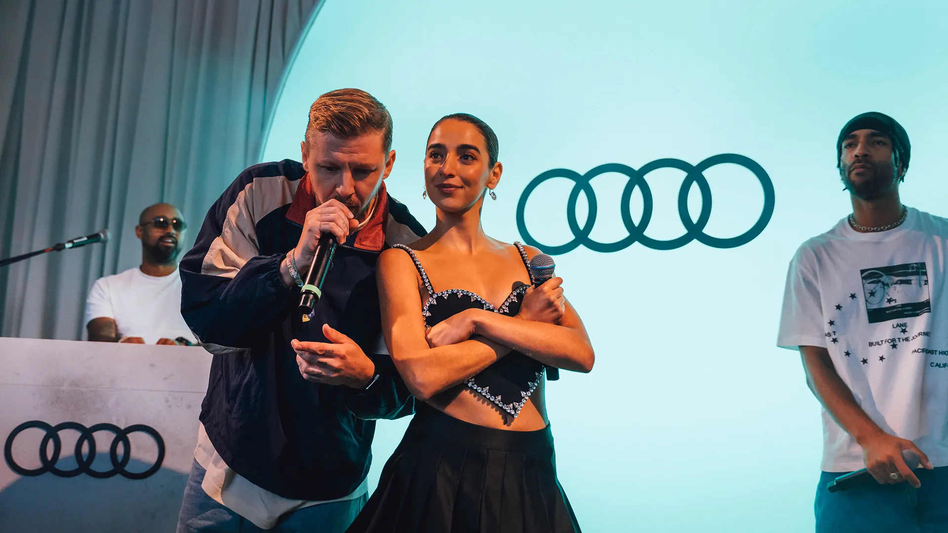 A male performer with a microphone leans in beside a woman with crossed arms on stage, with another performer nearby and the Audi logo projected behind them. A male performer with a microphone leans in beside a woman with crossed arms on stage, with another performer nearby and the Audi logo projected behind them.