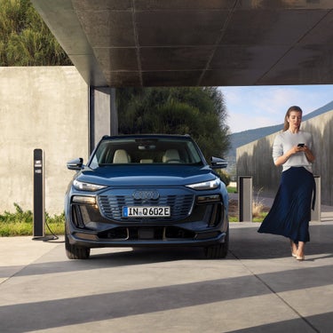 Ein blauer Audi Q6 SUV e-tron steht unter einem Carport, eine Frau läuft mit Smartphone vorbei, daneben eine Ladesäule.