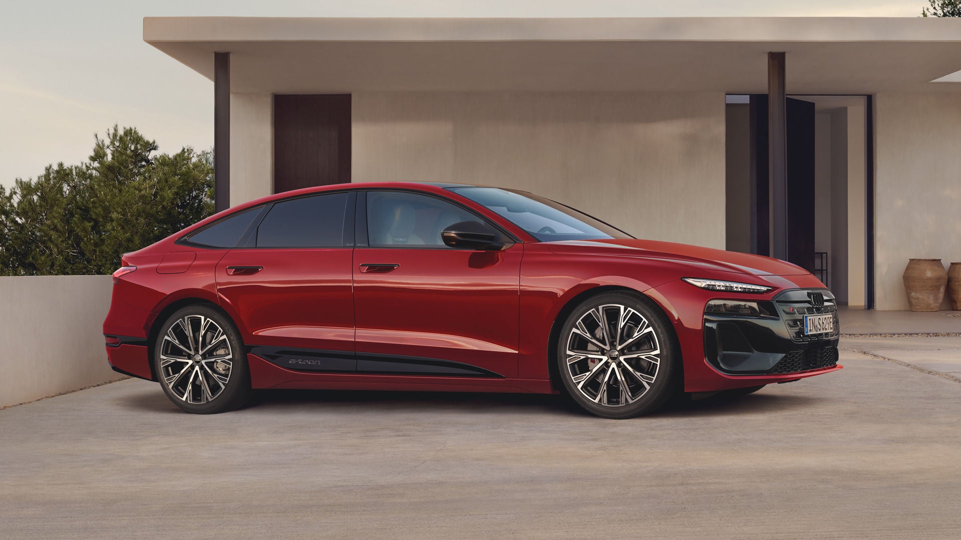 Der Audi S6 Sportback e-tron mit Individuallackierung Audi exclusive Granatrot Perleffekt, leicht von der Seite fotografiert, parkt vor einem modernen Wohngebäude.