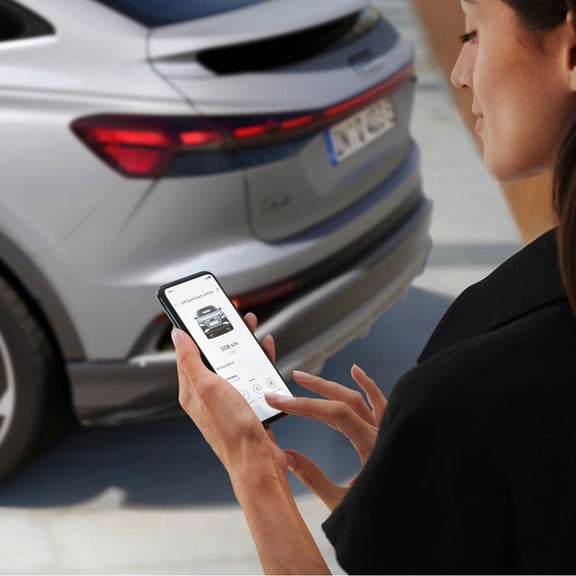A person using a smartphone to interact with a parked Audi Q4 Sportback e-tron nearby, showcasing modern technology and connectivity.