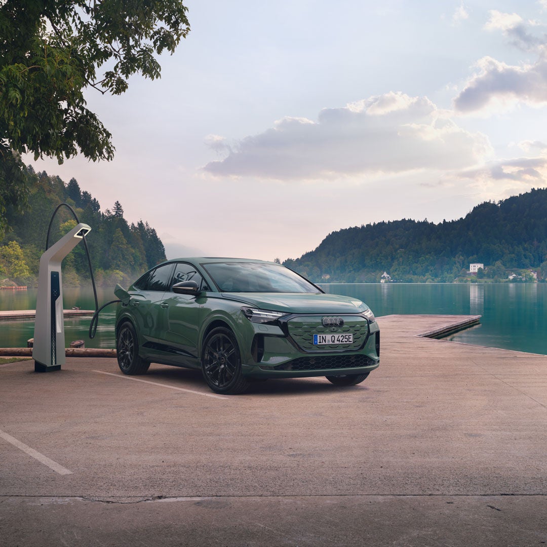 Audi Q4 Sportback e-tron verte en recharge sur une borne électrique au bord d’un lac, avec un ponton et des collines boisées en arrière-plan. 