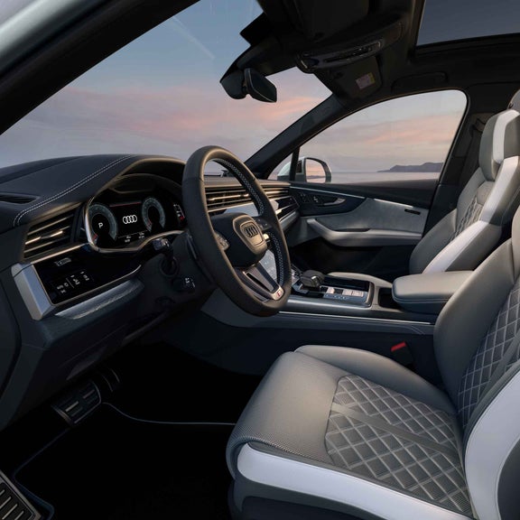Innenraum eines Audi Q7 SUV TSFI e mit schwarz Leder, Sportsitzen, digitalem Cockpit und Lenkrad, Blick auf die Fahrerseite geparkt vor einer Landschaft.