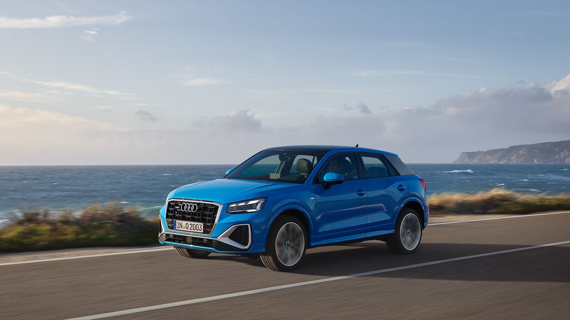 Blauer Audi Q2 fährt auf einer Küstenstraße mit Blick auf das Meer und eine felsige Landschaft im Hintergrund.