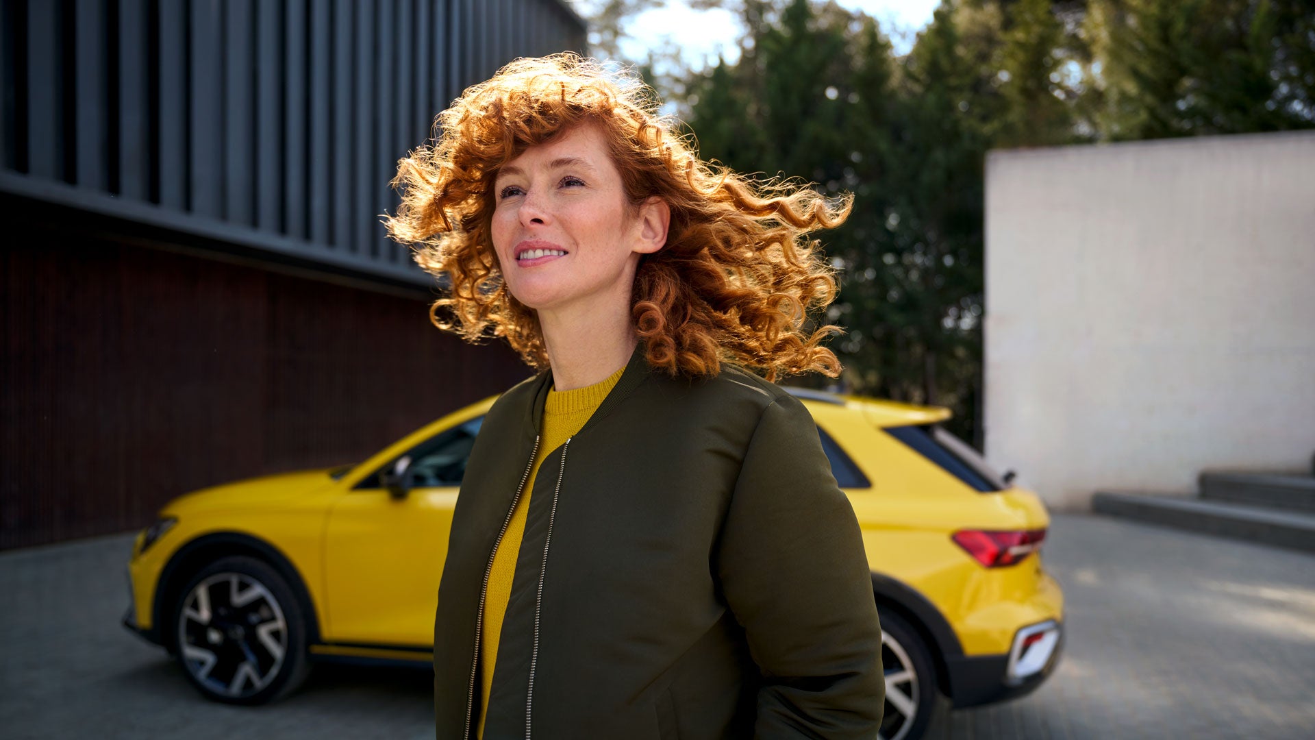 Eine Frau mit roten Locken steht lächelnd im Freien, im Hintergrund ein gelber Audi vor einem modernen Gebäude.
