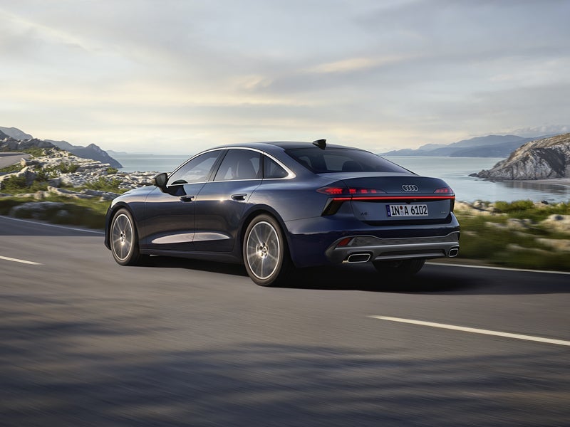 Die A6 Limousine in einer schrägen Heckperspektive, während sie auf einer kurvigen Straße entlang eines Fjords fährt.