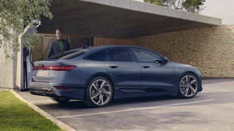 Der A6 Sportback e-tron steht rückwärts auf einem Parkplatz an einer Ladestation. Im Hintergrund ist ein modernes Gebäude zu sehen, sowie ein Mann im Anzug.