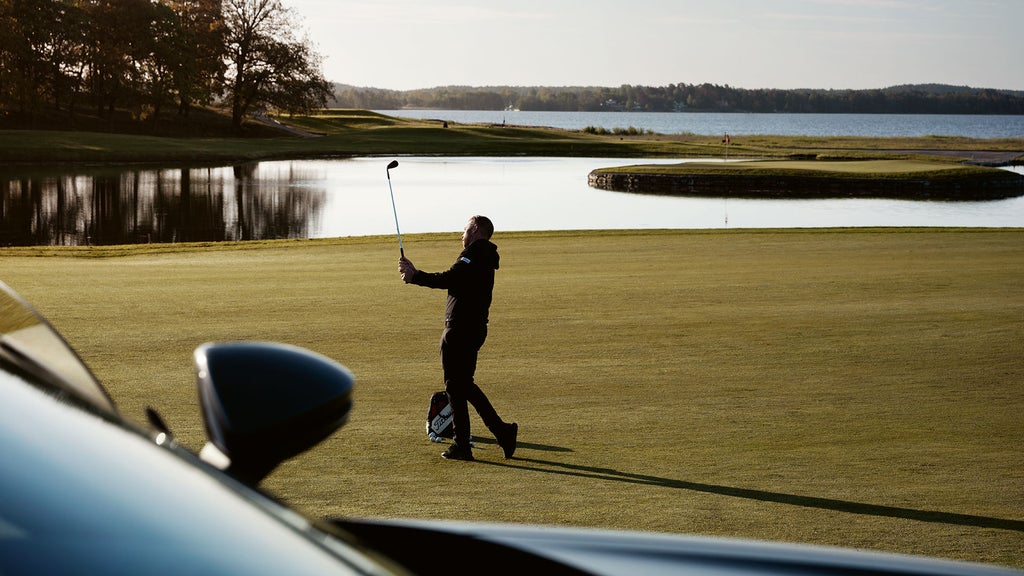 Vy över golfbana där Viktor svingar sin golfklubba. Sidospegeln på Viktors Audi syns i förgrunden.