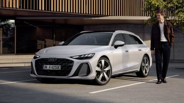 The A6 Avant e-hybrid stands at an angle in front of a large building with wood paneling in a parking lot. A middle-aged man stands next to the vehicle.