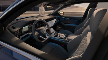 A view of the cockpit of the SQ8 SUV from the driver's side, with a futuristic, curved building in the background. A fashionably dressed young woman stands in front of the vehicle.
