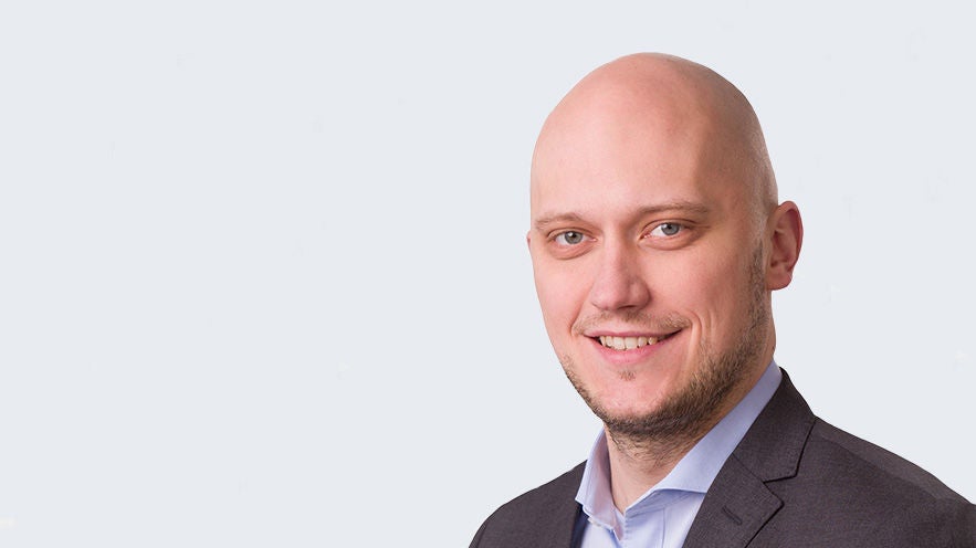 Andreas Büse: Porträt jüngerer, lächelnder Mann mit leichtem Bart in Anzug vor grauer Wand.