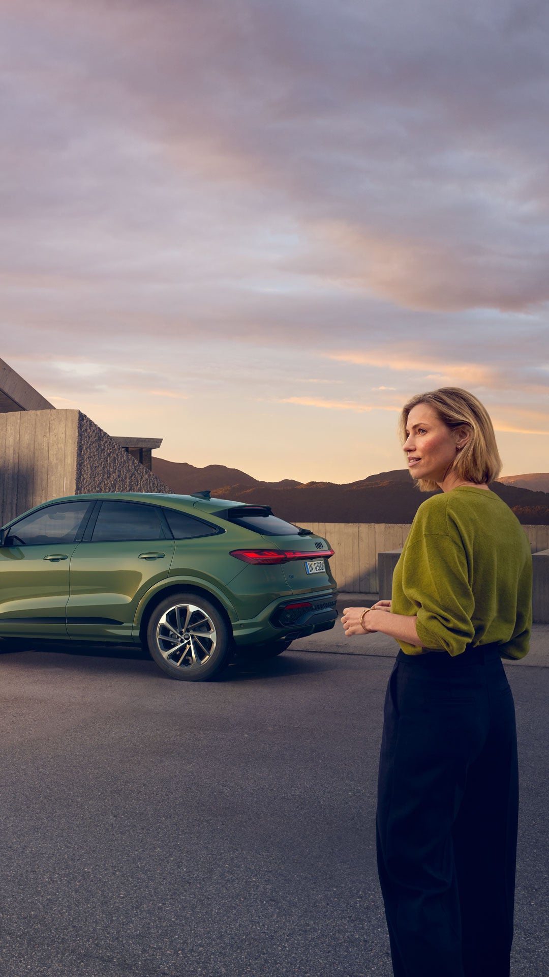 Eine Frau steht vor einem modernen Gebäude, im Hintergrund ein grüner Audi SUV und eine Berglandschaft bei Sonnenuntergang.