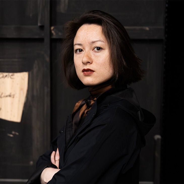 Frau mit kurzen dunklen Haaren blickt ernst in die Kamera, schwarzer Hintergrund und dunkle Kleidung.