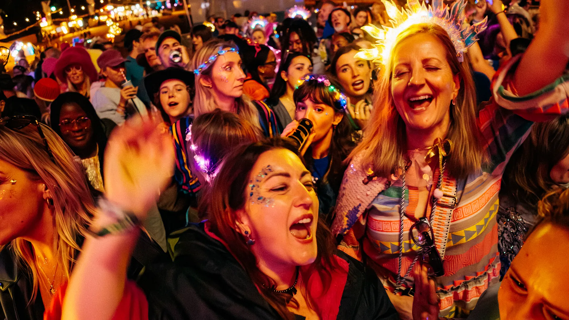 A joyful nighttime crowd wearing light-up crowns and glittery face decorations sings and dances together at the festival. A joyful nighttime crowd wearing light-up crowns and glittery face decorations sings and dances together at the festival.