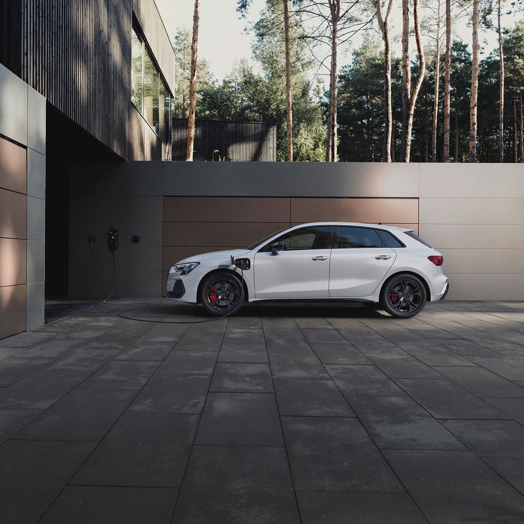 Audi A3 Sportback e-hybrid blanche en recharge dans l’allée d’une maison moderne, entourée d’une forêt de pins et d’une architecture contemporaine. 