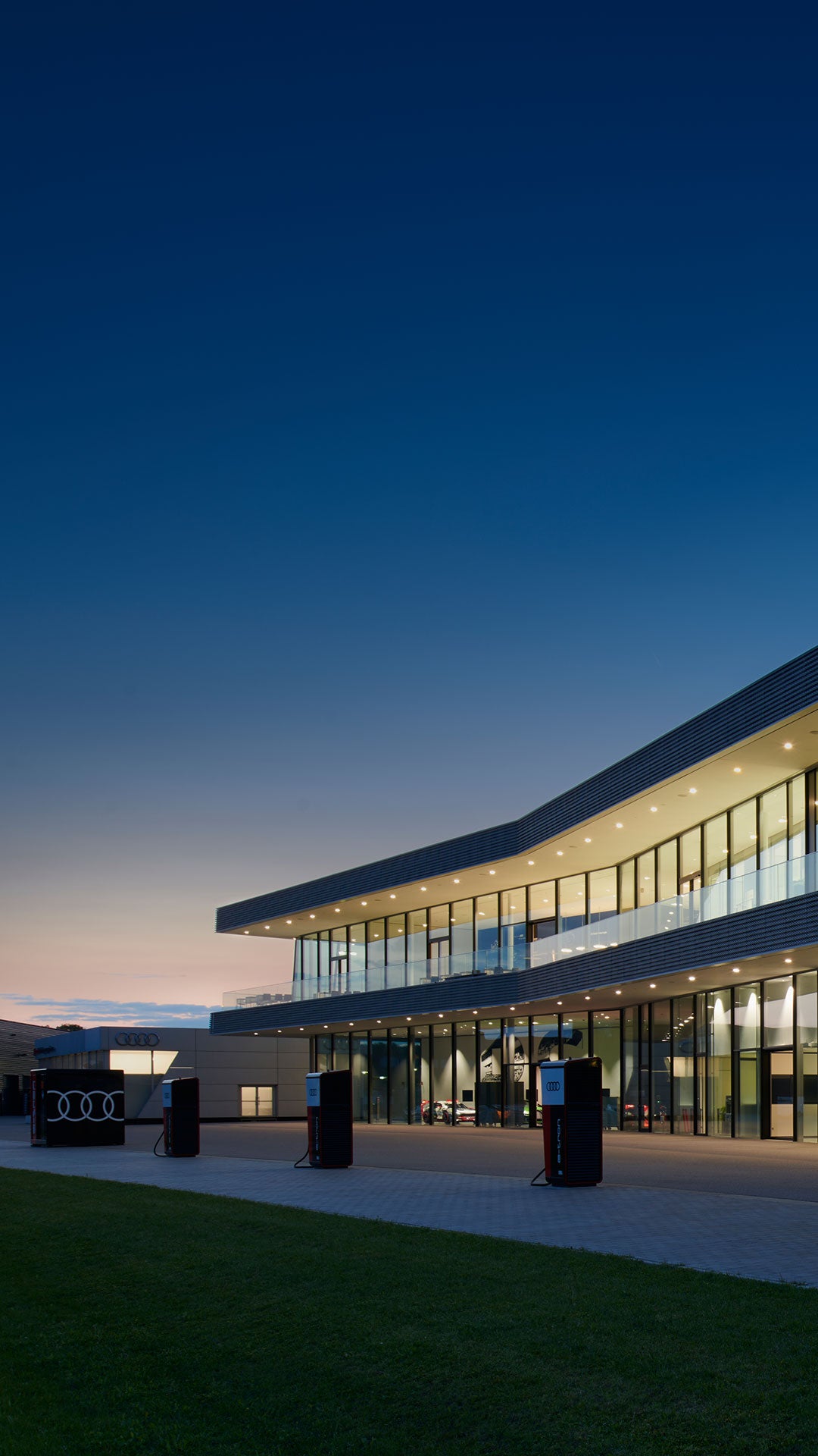 Glasfassade eines modernen Audi Gebäudes bei Abenddämmerung, im Vordergrund E-Ladesäulen.