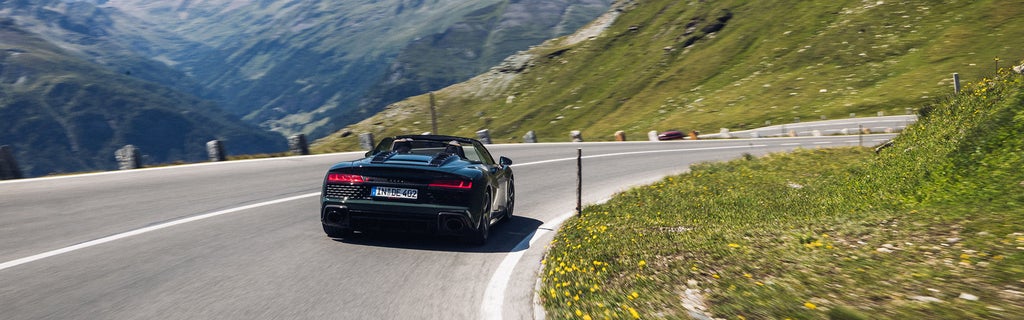 Ein schwarzer Audi R8 Spyder V10 performance quattro mit offenem Verdeck (Heckansicht) fährt durch eine scharfe Kurve in alpiner Landschaft bei klarem Himmel.