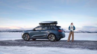 Dunkelblauer Audi Q6 e-tron mit Dachbox steht bei Sonnenaufgang vor modernem Haus, Frau blickt aus verglaster Terrasse.