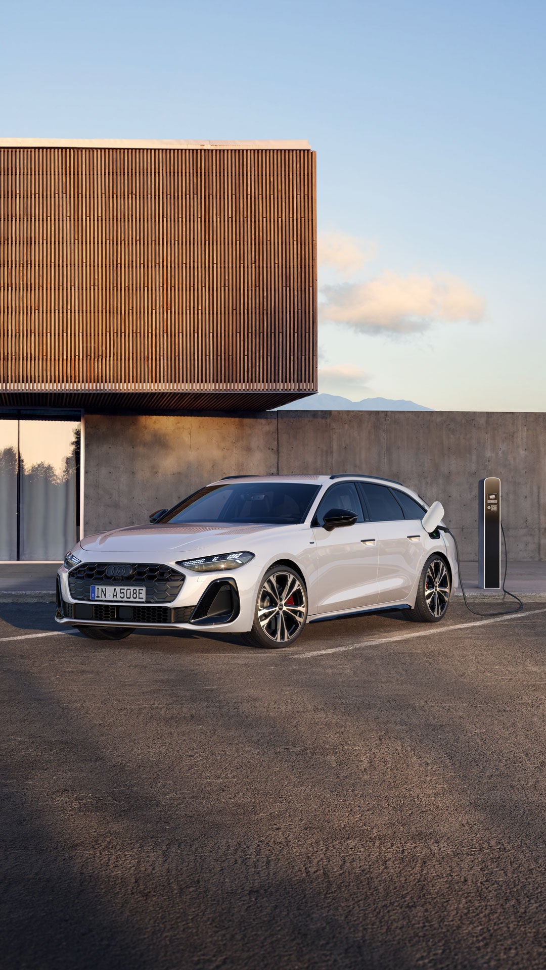 Weißer Audi A6 Avant e-tron lädt an einer Ladesäule auf einem Parkplatz vor Betonwand.
