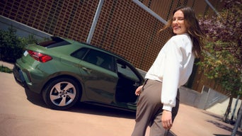 Lächelnde Frau blickt zurück, während sie neben einem grünen Audi A3 mit geöffneter Tür auf einem sonnigen Gehweg steht.