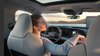 Frau sitzt entspannt mit geschlossenen Augen im Audi SUV Q6 e-tron, mit Blick auf Meer und Sonnenuntergang durch die Frontscheibe.