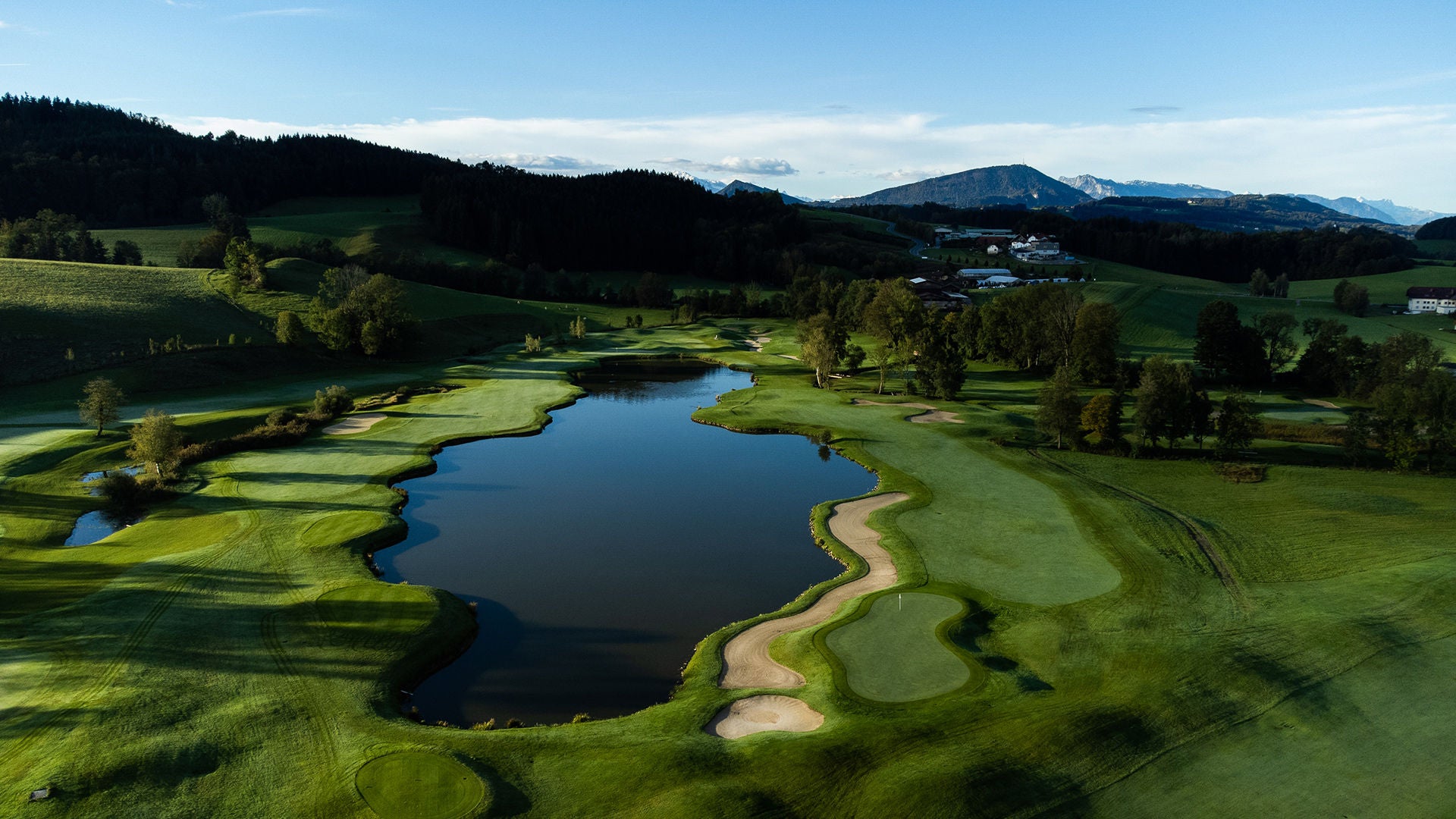 A sprawling golf course with a lake, sand bunkers, rolling hills, and blue skies.