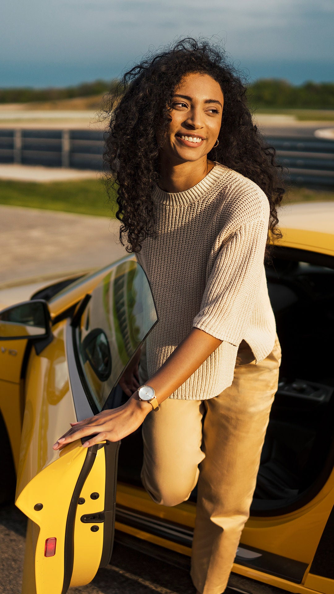 Farbige Frau mit lockigem Haar steigt lächelnd aus gelben Audi Modell bei sonnigem Wetter und blauem Himmel.