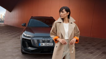 Eine Frau im beigen Mantel steht vor einem dunkelroten Audi SQ6 e-tron, im Hintergrund eine moderne rote Gebäudewand.