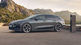 Der A6 Avant e-tron steht seitlich auf einem Kiesparkplatz vor einem See und ist dabei an eine Ladestation angeschlossen. Im Hintergrund sind von Bäumen bewachsene Berge zu sehen.