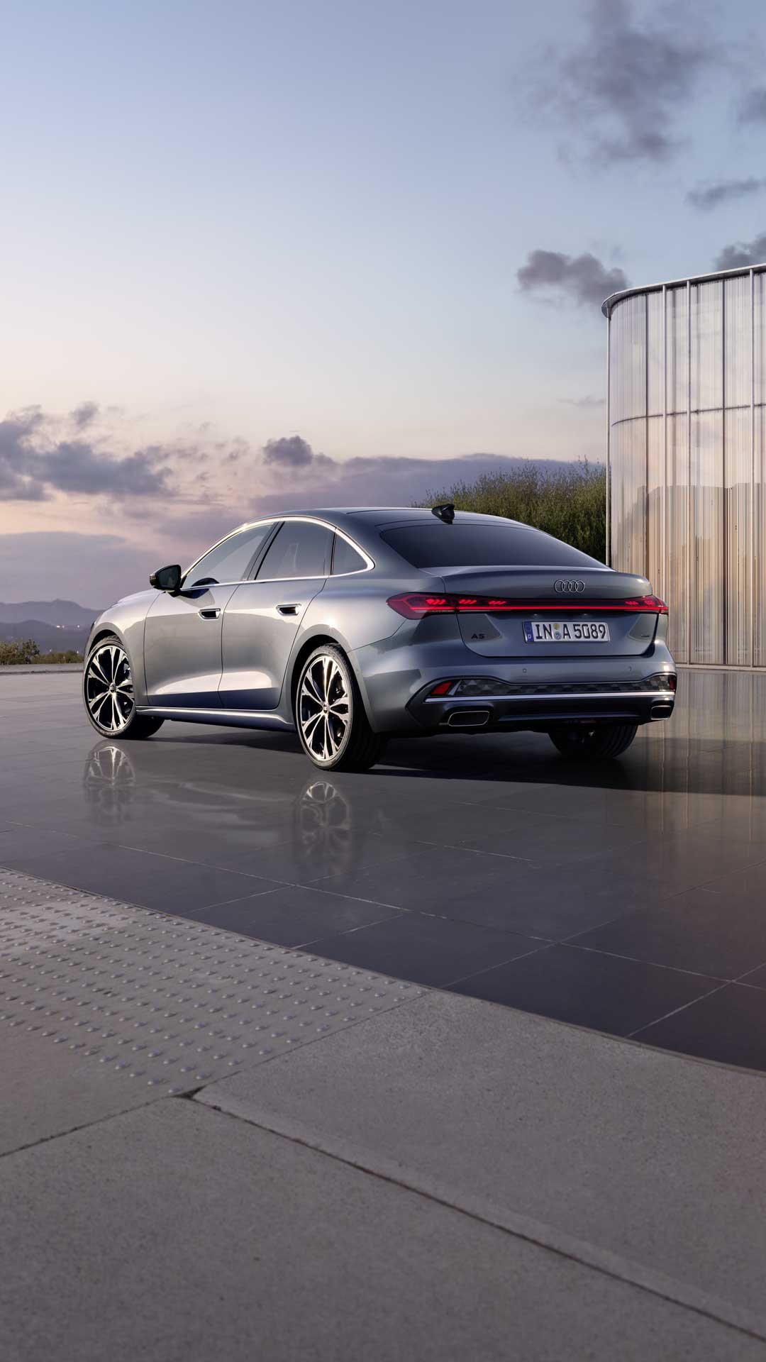 Die A5 Limousine im schrägen Heckprofil. Das Fahrzeug spiegelt sich auf dem glatten Untergrund. Im Hintergrund sind die Konturen von Bergen bei Dämmerung zu sehen.