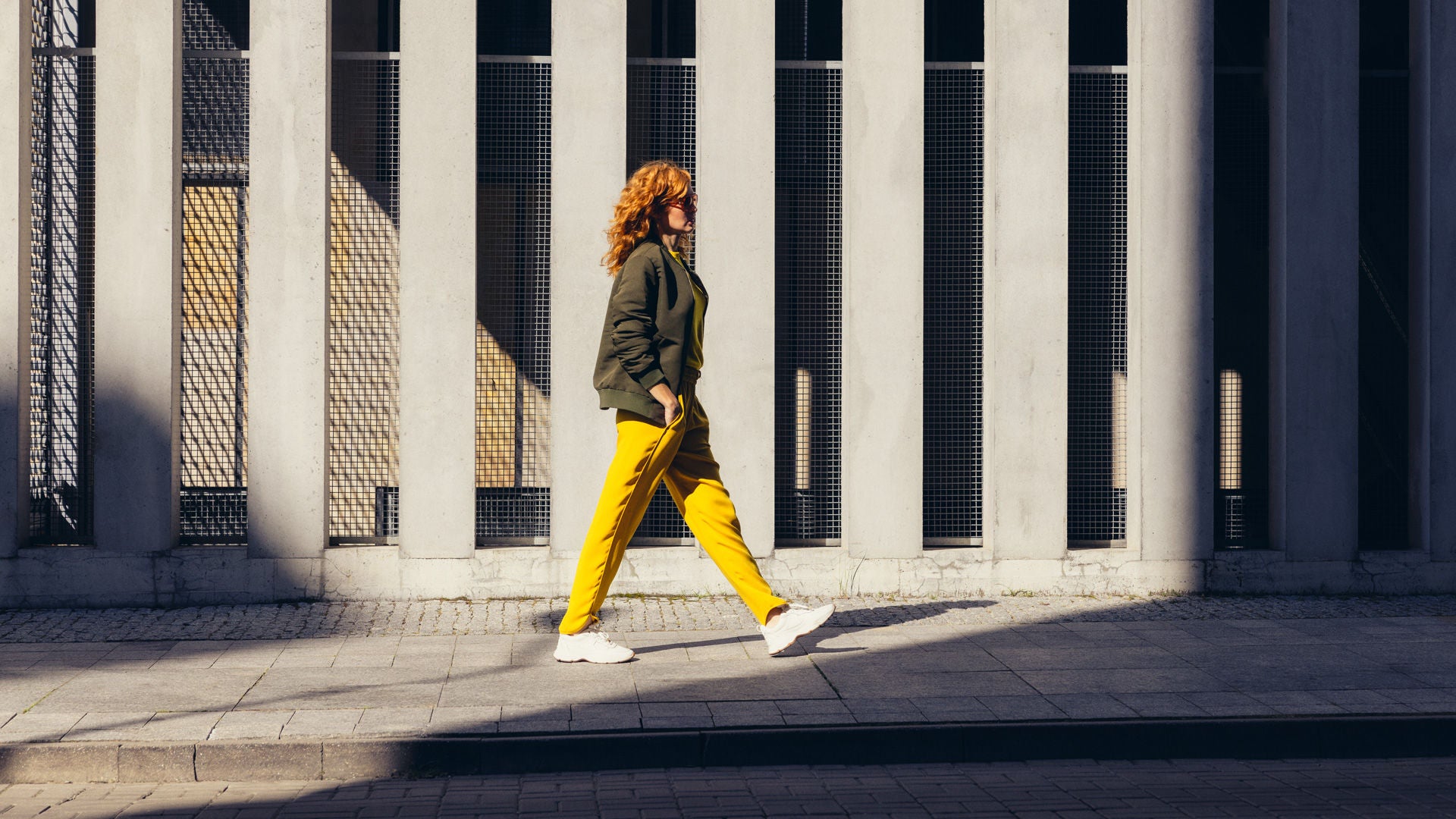 Eine Person mit roten, lockigen Haaren geht seitlich an einer modernen Betonfassade mit vertikalen Streben vorbei; sie trägt eine grüne Jacke, gelbe Hose und weiße Sneaker, während Sonnenlicht starke Schatten auf den Gehweg wirft.