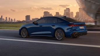 Eine blaue Audi S5 Limousine fährt bei Sonnenuntergang auf einer Straße mit Stadtpanorama im Hintergrund.
