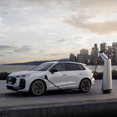 Weißer Audi Q3 SUV e-hybrid an einer silbernen Ladesäule vor einer Großstadtkulisse im Hintergrund.