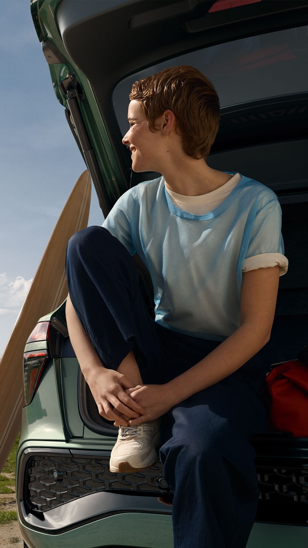 Une jeune femme est assise dans le coffre d'une Audi Q3 verte et regarde en direction de la plage et de la mer. Une planche de surf est appuyée contre la voiture.