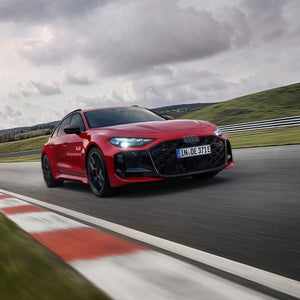 Ein roter Audi RS 5 Avant fährt auf einer Rennstrecke. Im Vordergrund sind rot-weiße Curbs zu erkennen, im Hintergrund eine grüne Hügellandschaft.