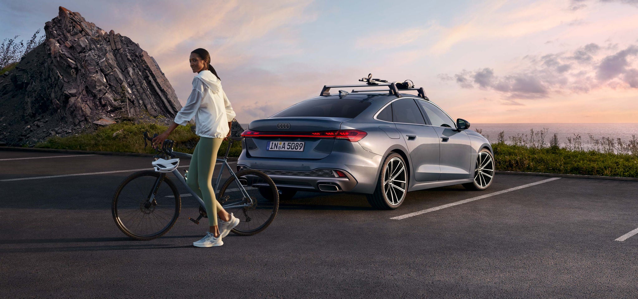 The A5 Saloon with a sloping rear profile. The vehicle is parked in a parking lot near a mountain. There is a bike rack on the roof. Behind the vehicle, a young woman is pushing a bicycle.