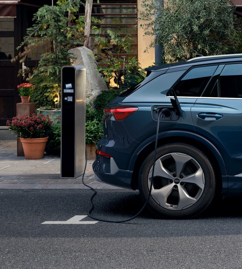 Nahaufnahme des rechten Hecks des Q4 SUV e-tron, parkend vor einem Gebäude, Pflanztöpfe sind vor dem Gebäude zu sehen. Das Fahrzeug ist an eine öffentliche Ladesäule angeschlossen.&nbsp;