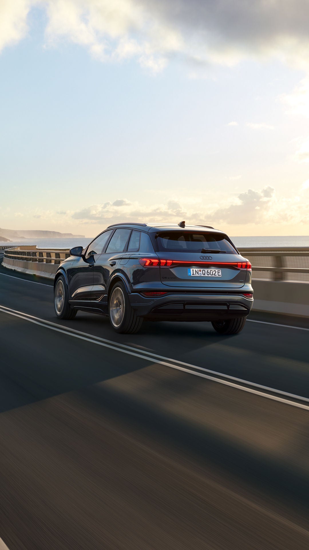 Schräge Heckaufnahme des Q6 SUV e-tron, welche über eine kurvige Straße am Meer fährt. Im Hintergrund sind Berge zu sehen.