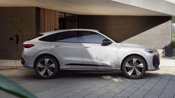 The Q5 Sportback e-hybrid is shown in side profile in front of a modern house. The vehicle is connected to a charging station mounted on the wall of the house.